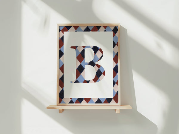 A framed art print featuring a large letter 'B' filled with a geometric diamond pattern, displayed on a light wooden shelf against a white wall with soft window shadows.