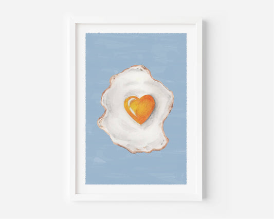 A framed art print featuring a painted fried egg with a bright orange heart-shaped yolk on a light blue background, displayed in a white frame against a white wall.