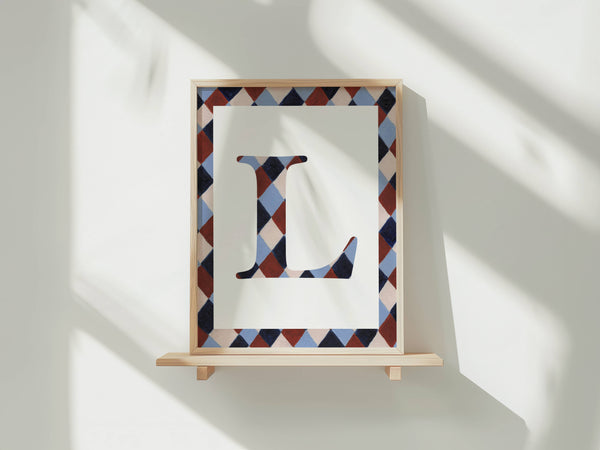 A framed letter monogram print featuring a large capital letter 'L' in a geometric diamond pattern, mounted on a light wood shelf against a white wall with soft window shadows.