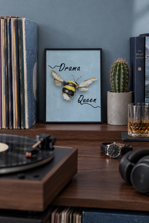 A framed art print featuring a watercolor bumblebee with the words 'Drama Queen' on a light blue background, styled on a wooden shelf next to vinyl records, a cactus, a glass of whiskey, and headphones.