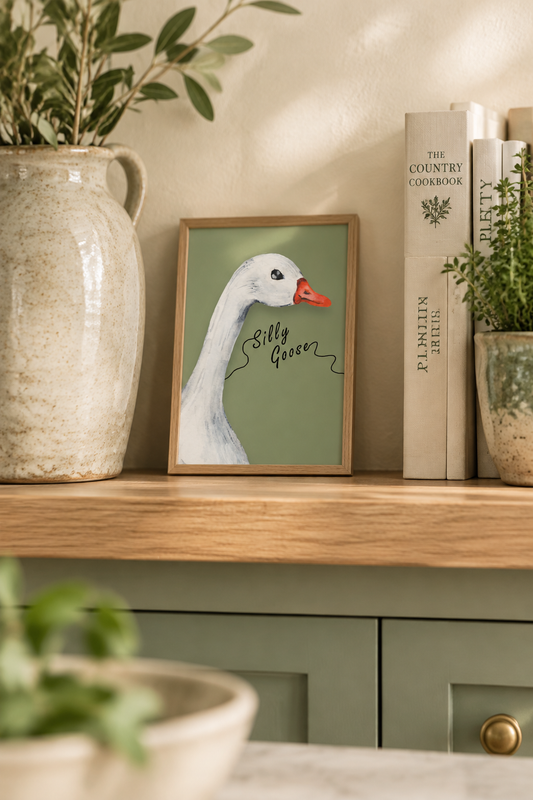 Silly Goose in a country kitchen: oak frame propped on a shelf with a stoneware jug, cookbooks and a potted herb for scale, in cream and sage tones that match the print's background.
