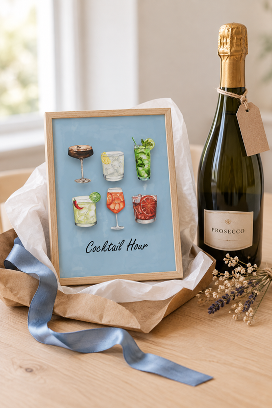 A framed art print titled 'Cocktail Hour' featuring six colorful cocktail illustrations on a light blue background, styled on a wooden table next to a Prosecco bottle and wrapped in tissue paper.