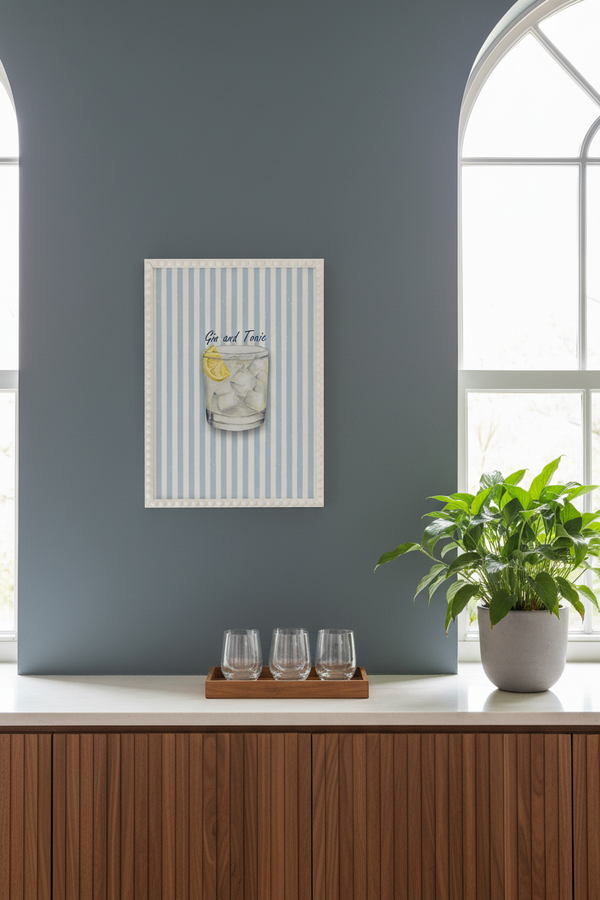 A framed art print featuring a cocktail glass illustration with the words 'Gin and Tonic" and light blue and white vertical stripes, displayed on a dark blue-gray wall above a wooden counter with wine glasses and a potted plant.