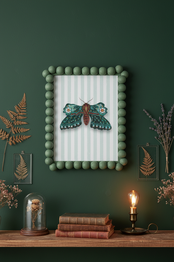 A framed wall art piece featuring a detailed green moth on a white striped background, surrounded by a green pom-pom border, displayed on a dark green wall above a wooden shelf with books, a candle, and dried ferns.