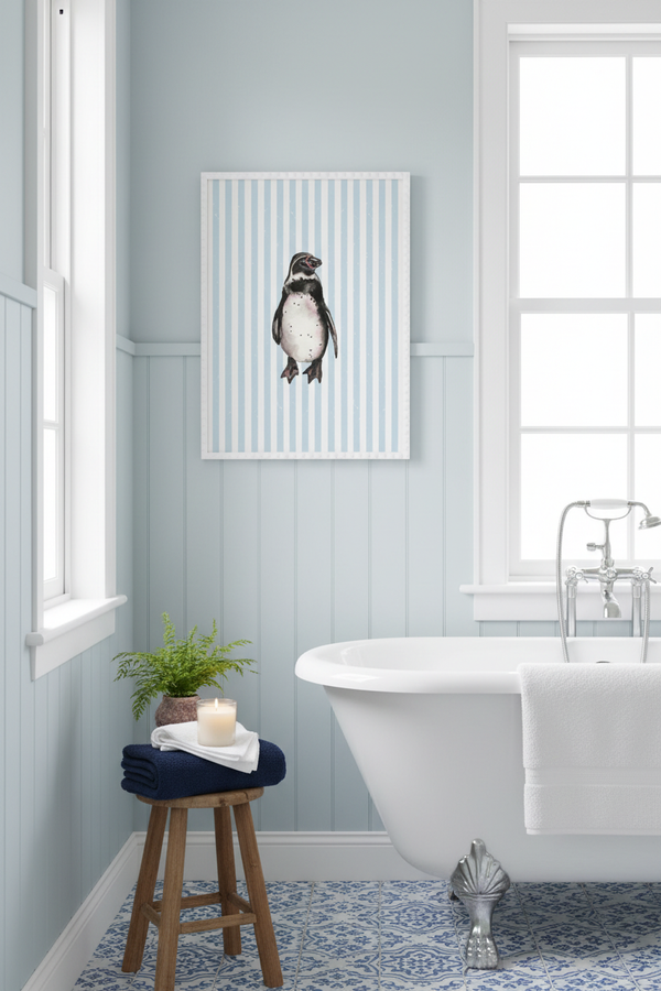 A framed wall art print featuring a penguin on vertical light blue and white stripes, displayed in a bright bathroom above a white clawfoot tub and next to a small wooden stool with a plant and candle.
