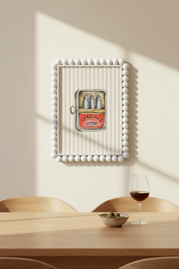 A framed wall art piece hangs on a beige wall above a wooden dining table. The artwork features a white beaded border, vertical cream stripes, and a vintage-style can-shaped illustration with colorful fish and a red label.