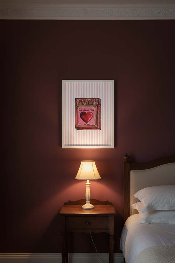 A framed art print featuring a pink heart and the words 'I Love You' hanging on a dark burgundy wall above a wooden nightstand with a lit table lamp and a bed with white linens.