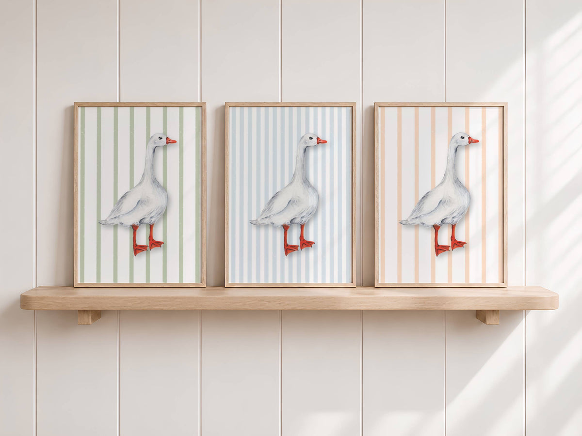 Three framed vertical art prints of a white goose with red-orange legs and beak, set against striped backgrounds in soft green, blue, and peach tones, displayed on a light wood floating shelf against a white paneled wall.