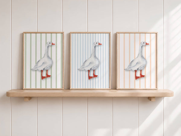 Three framed vertical art prints of a white goose with red-orange legs and beak, set against striped backgrounds in soft green, blue, and peach tones, displayed on a light wood floating shelf against a white paneled wall.