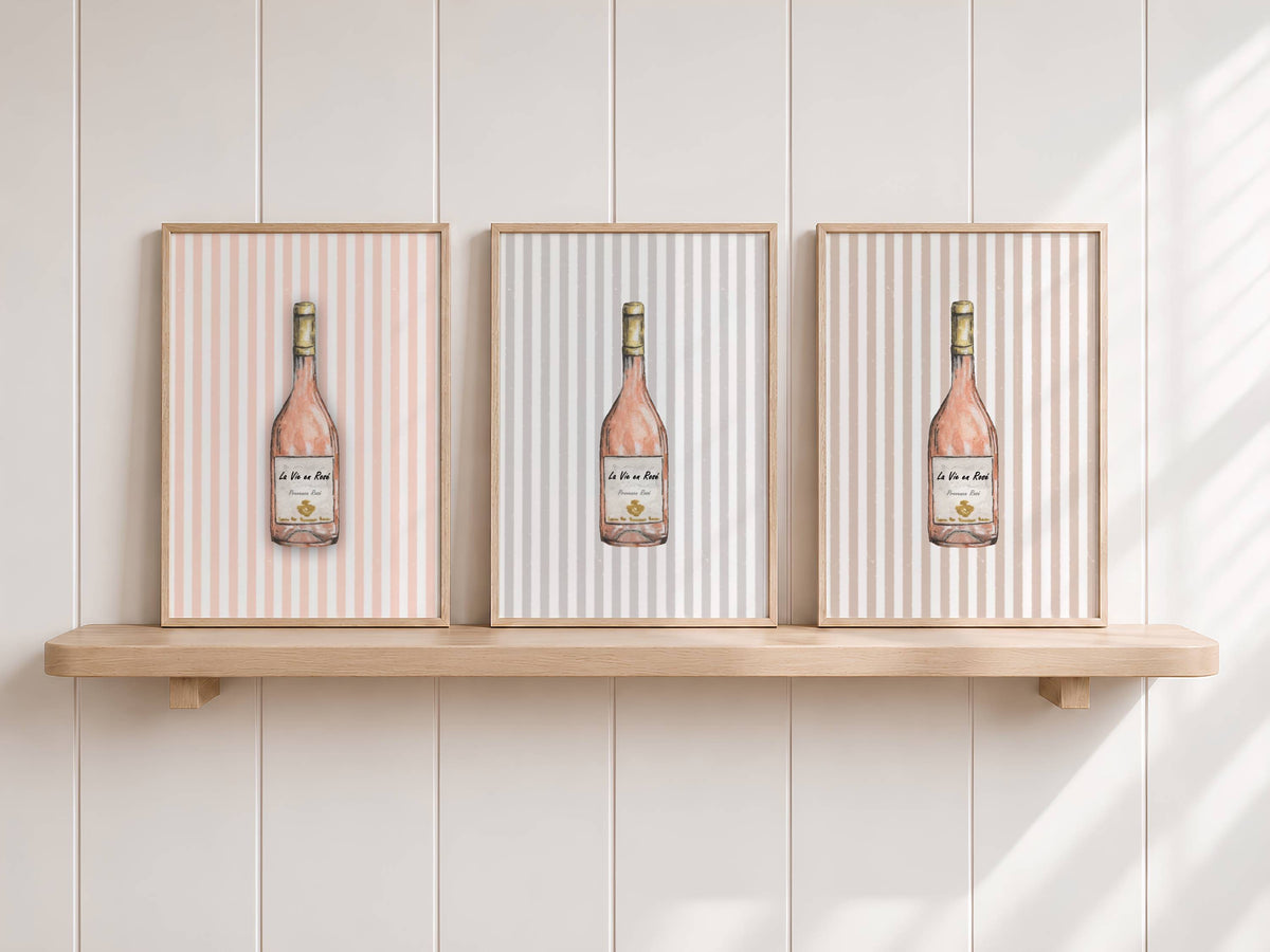 Three framed wall art prints with vertical pink and white stripes and a central wine bottle illustration, displayed on a light wood shelf against a white paneled wall.