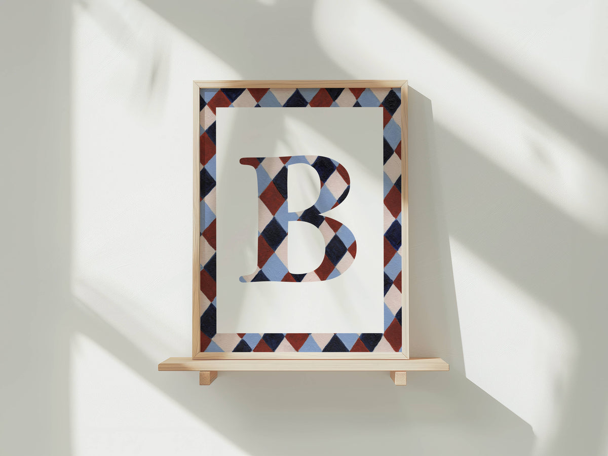 A framed art print featuring a large letter 'B' filled with a geometric diamond pattern, displayed on a light wooden shelf against a white wall with soft window shadows.