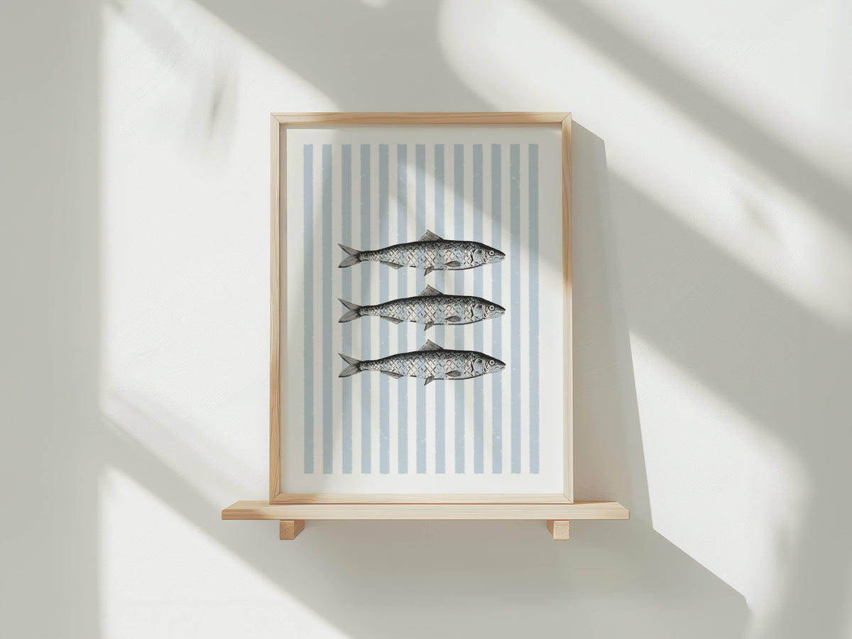 Framed wall art featuring three striped fish on a white background with vertical light blue stripes, displayed on a light wood shelf against a white wall with soft window shadows.
