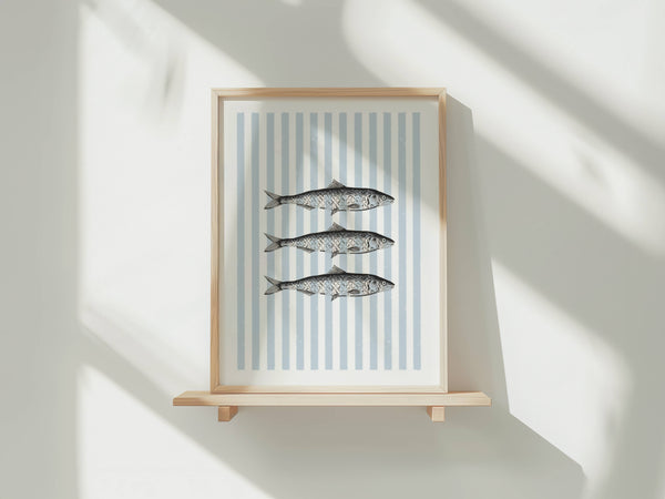 Framed wall art featuring three striped fish on a white background with vertical light blue stripes, displayed on a light wood shelf against a white wall with soft window shadows.