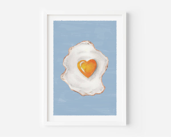 A framed art print featuring a painted fried egg with a bright orange heart-shaped yolk on a light blue background, displayed in a white frame against a white wall.