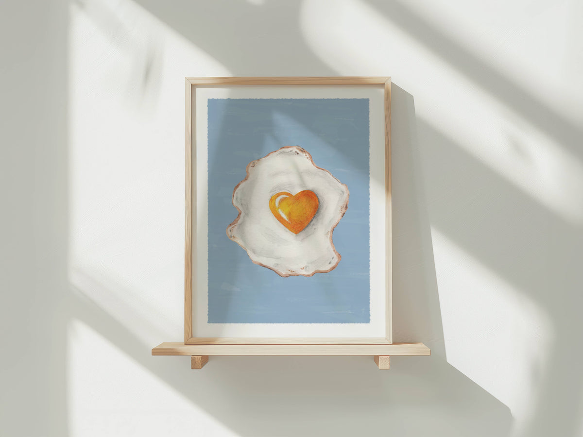 A framed art print featuring a stylized fried egg with a heart-shaped yolk on a light blue background, displayed on a wooden shelf against a white wall with soft window shadows.