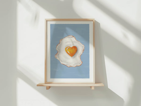 A framed art print featuring a stylized fried egg with a heart-shaped yolk on a light blue background, displayed on a wooden shelf against a white wall with soft window shadows.