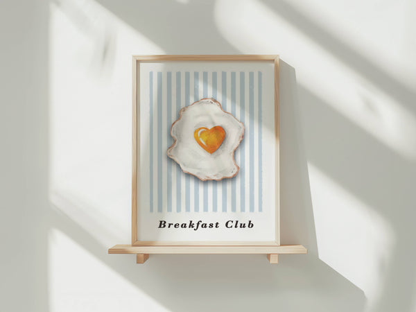 A framed art print featuring a stylized fried egg with a heart-shaped yolk, displayed on a light wooden stand against a white wall with soft window shadows.