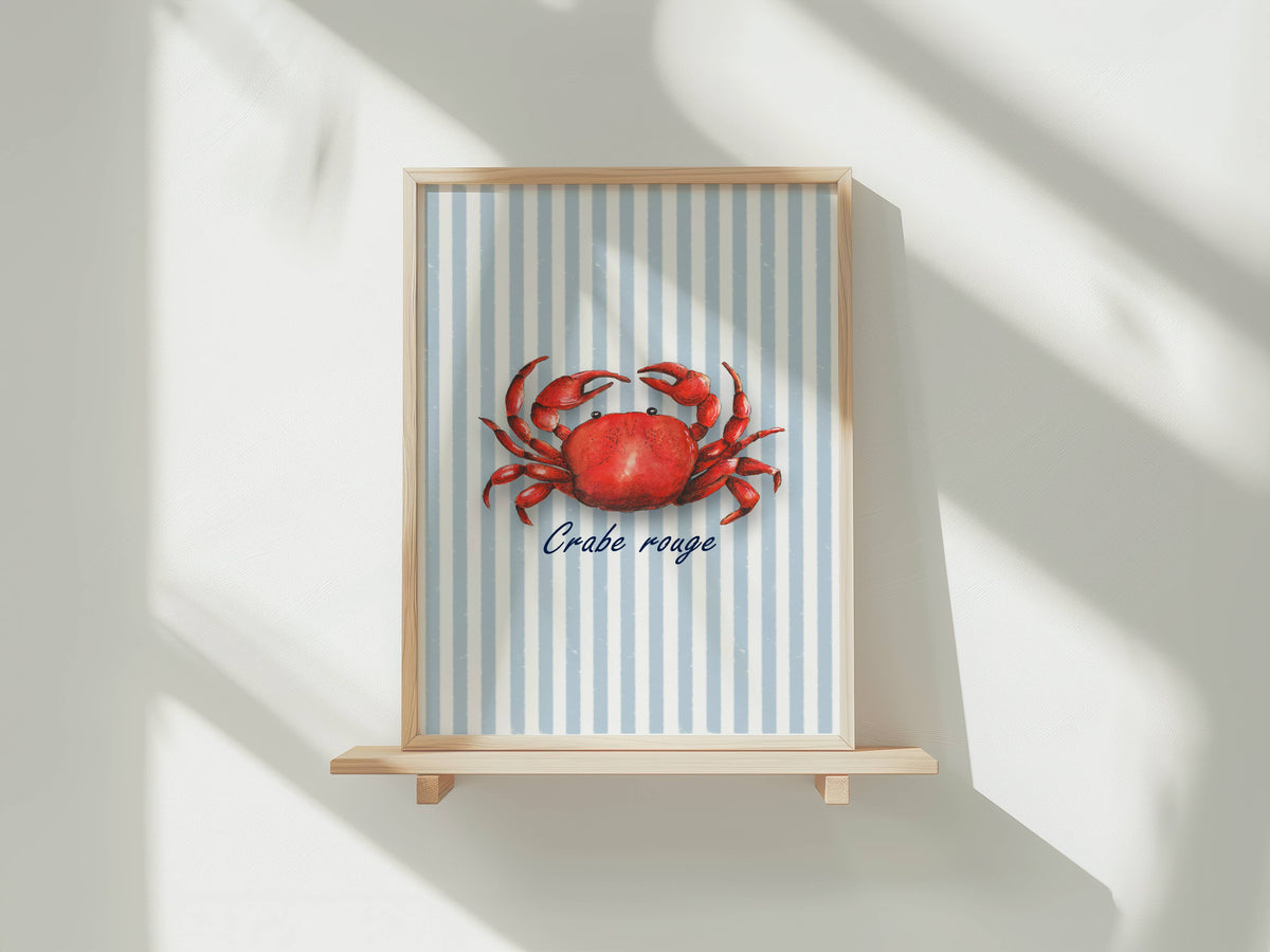 A framed art print featuring a red crab on a light blue and white striped background with the French text 'Crabe rouge' below it, displayed on a light wooden shelf against a white wall with soft diagonal shadows.