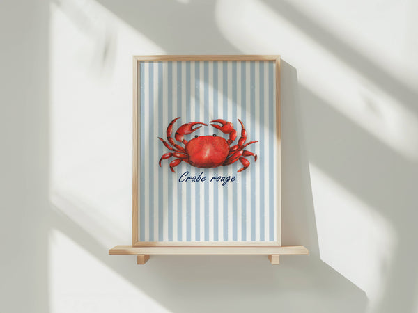 A framed art print featuring a red crab on a light blue and white striped background with the French text 'Crabe rouge' below it, displayed on a light wooden shelf against a white wall with soft diagonal shadows.