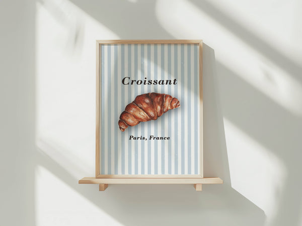 A framed art print featuring a croissant on a light blue and white striped background with the text 'Croissant' and 'Paris, France', displayed on a wooden shelf against a white wall with soft window shadows.