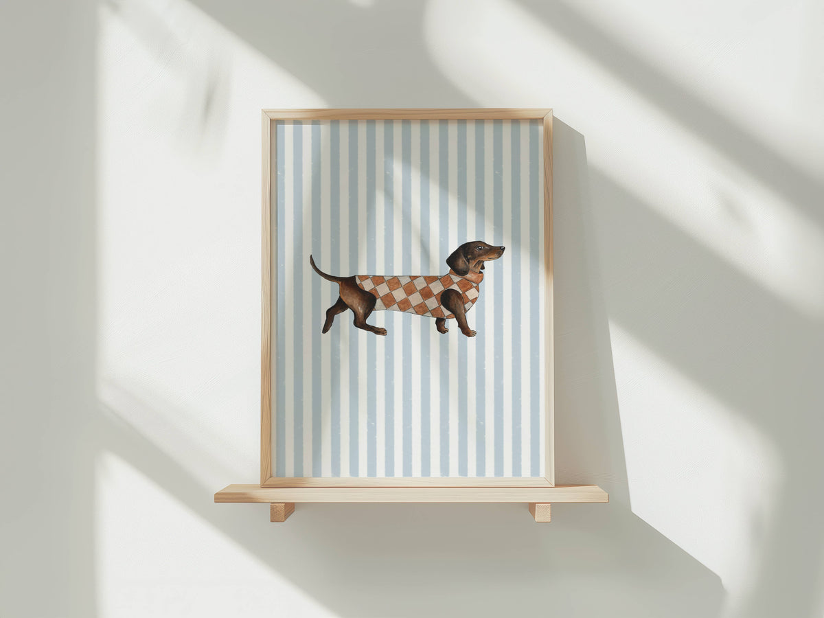 A framed art print featuring a dachshund wearing a brown and white argyle sweater, set against a background of light blue and white vertical stripes. The frame is light wood and displayed on a matching wooden shelf against a white wall with soft, diagonal shadow patterns.