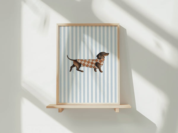 A framed art print featuring a dachshund wearing a brown and white argyle sweater, set against a background of light blue and white vertical stripes. The frame is light wood and displayed on a matching wooden shelf against a white wall with soft, diagonal shadow patterns.