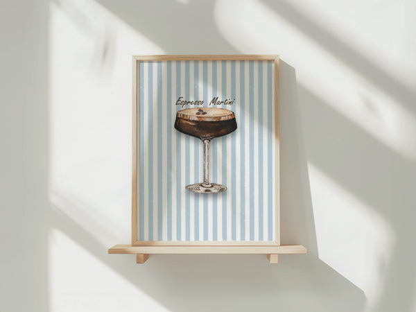 A framed art print featuring a watercolour illustration of an Espresso Martini cocktail in a coupe glass with coffee beans on top, set against a background of vertical light blue and white stripes. The print is shown in a light wood frame displayed on a wooden shelf against a white wall with soft window shadows.