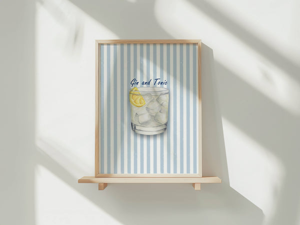 A framed art print featuring a watercolor illustration of a Gin and Tonic cocktail with a lemon slice, set against a light blue and white striped background. The print is housed in a light natural wood frame and displayed on a wooden shelf against a white wall with soft, diagonal shadows.