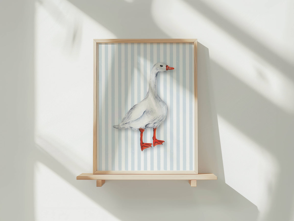 A framed art print featuring a white goose with red legs and beak, set against a light blue and white striped background, displayed on a light wood shelf against a white wall with soft window shadows.
