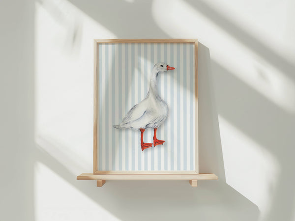 A framed art print featuring a white goose with red legs and beak, set against a light blue and white striped background, displayed on a light wood shelf against a white wall with soft window shadows.