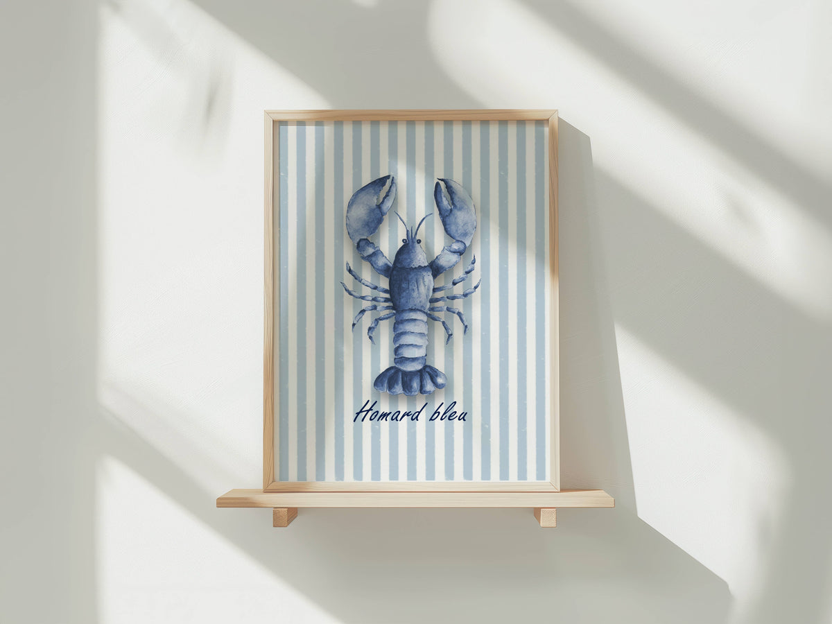 A framed art print featuring a blue lobster illustration on a light blue and white striped background, with the French text 'Homard bleu' below. The frame is mounted on a light wood shelf against a white wall with soft diagonal shadows.