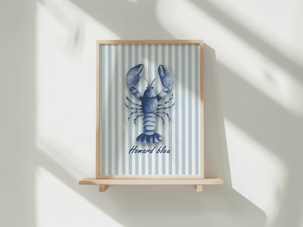 A framed art print featuring a blue lobster illustration on a light blue and white striped background, with the French text 'Homard bleu' below. The frame is mounted on a light wood shelf against a white wall with soft diagonal shadows.