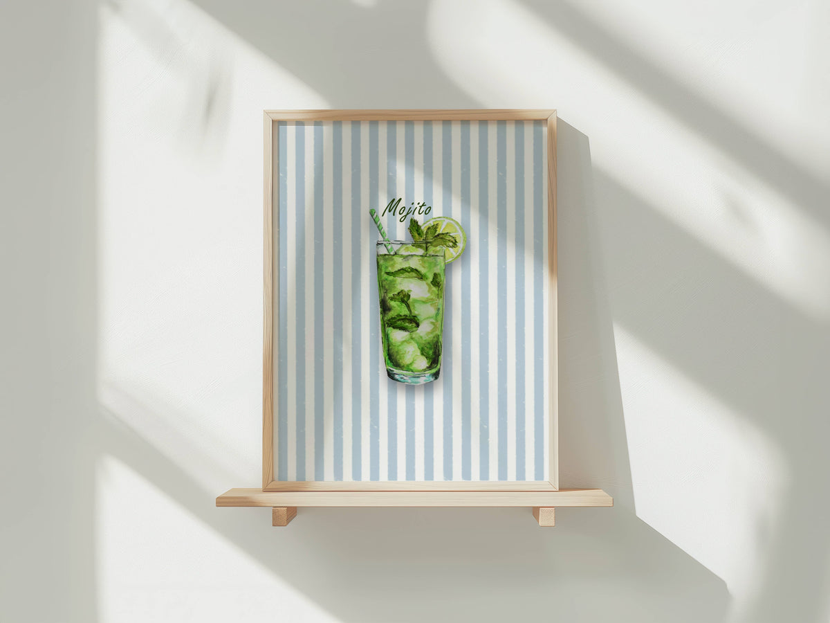 A framed art print featuring a watercolour illustration of a mojito cocktail in a tall glass with mint leaves, ice, a lime wheel and a striped straw, set against a background of vertical light blue and white stripes. The print is shown in a light wood frame displayed on a wooden shelf against a white wall with soft window shadows.