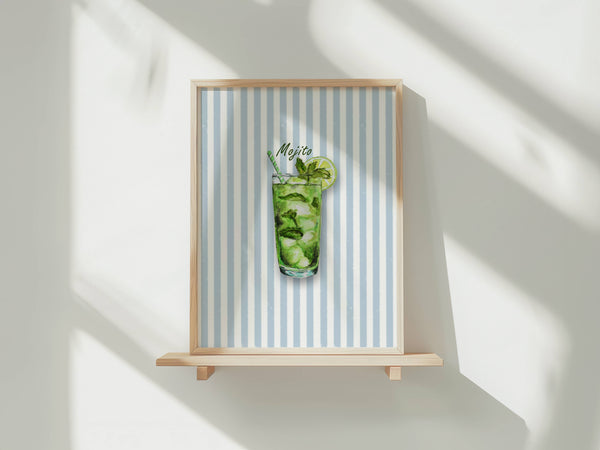 A framed art print featuring a watercolour illustration of a mojito cocktail in a tall glass with mint leaves, ice, a lime wheel and a striped straw, set against a background of vertical light blue and white stripes. The print is shown in a light wood frame displayed on a wooden shelf against a white wall with soft window shadows.