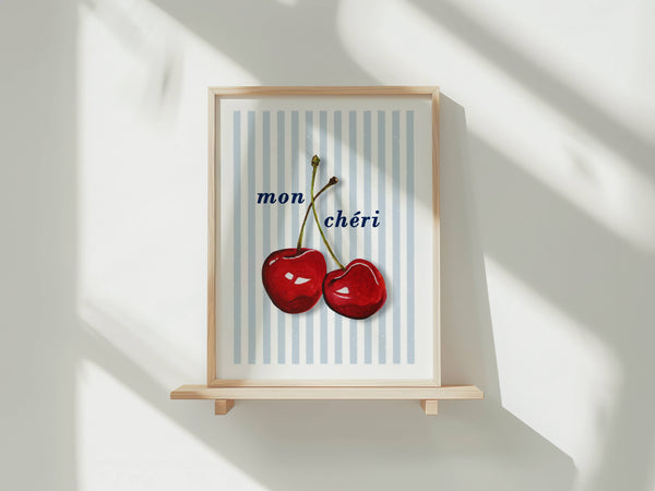 A framed art print featuring two red cherries on a light blue and white striped background with the French text 'mon chéri' in navy blue. The frame is light wood and displayed on a small wooden shelf against a white wall with soft diagonal shadows.