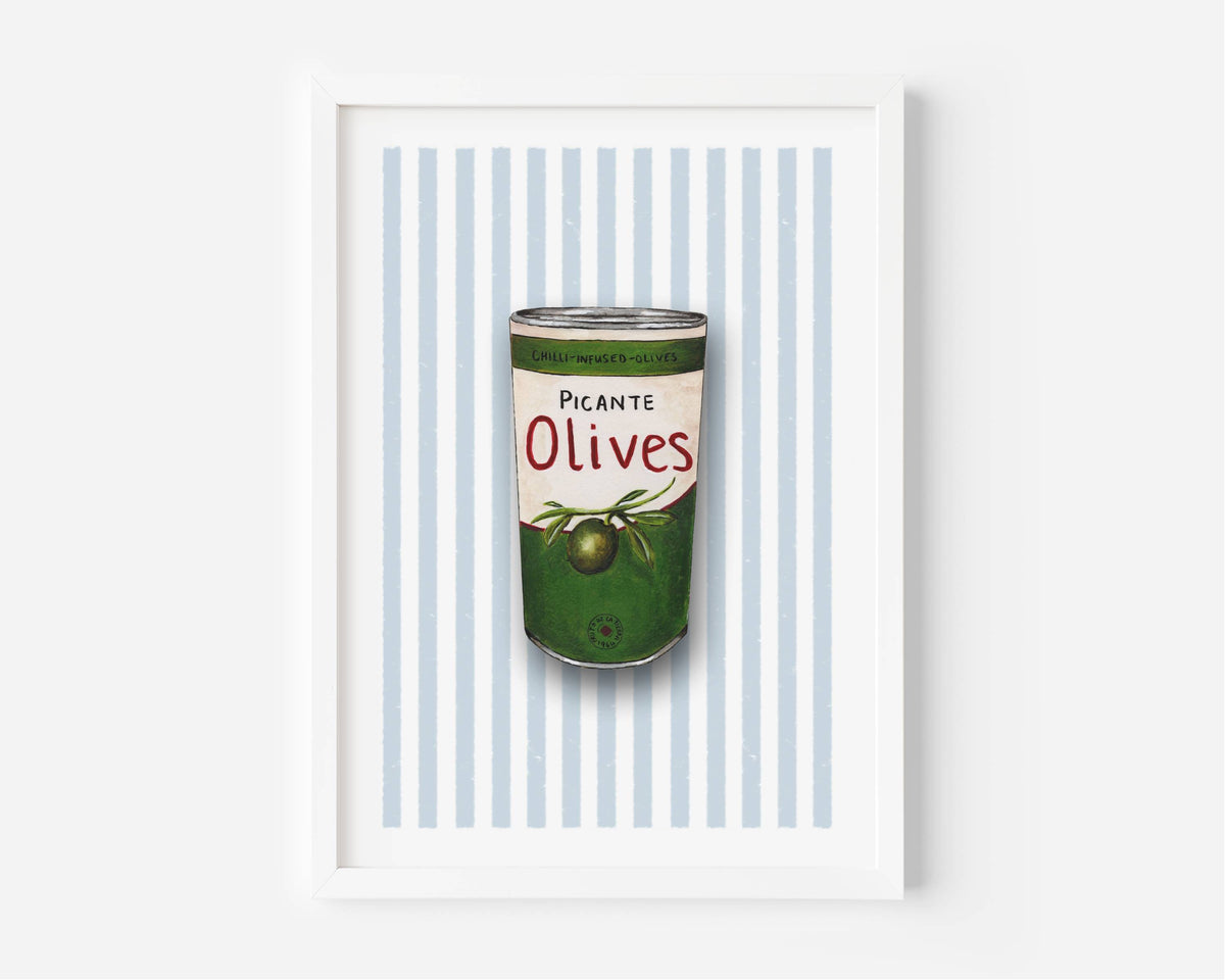 A framed art print featuring a vintage-style illustration of a green can of olives with the text 'Picante Olives' on a background of light blue and white vertical stripes, set against a clean white wall.
