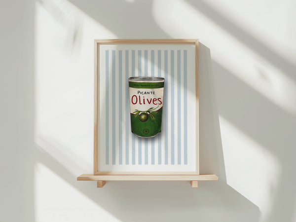 A framed art print displayed on a light wooden easel against a white wall with soft diagonal shadows. The print features a vintage-style illustration of a green can of 'Picante Olives' centered on a background of light blue and white vertical stripes.