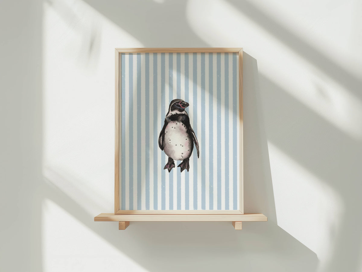 A framed art print featuring a penguin on a light blue and white striped background, displayed on a light wood floating shelf against a white wall with soft window shadows.
