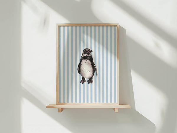 A framed art print featuring a penguin on a light blue and white striped background, displayed on a light wood floating shelf against a white wall with soft window shadows.