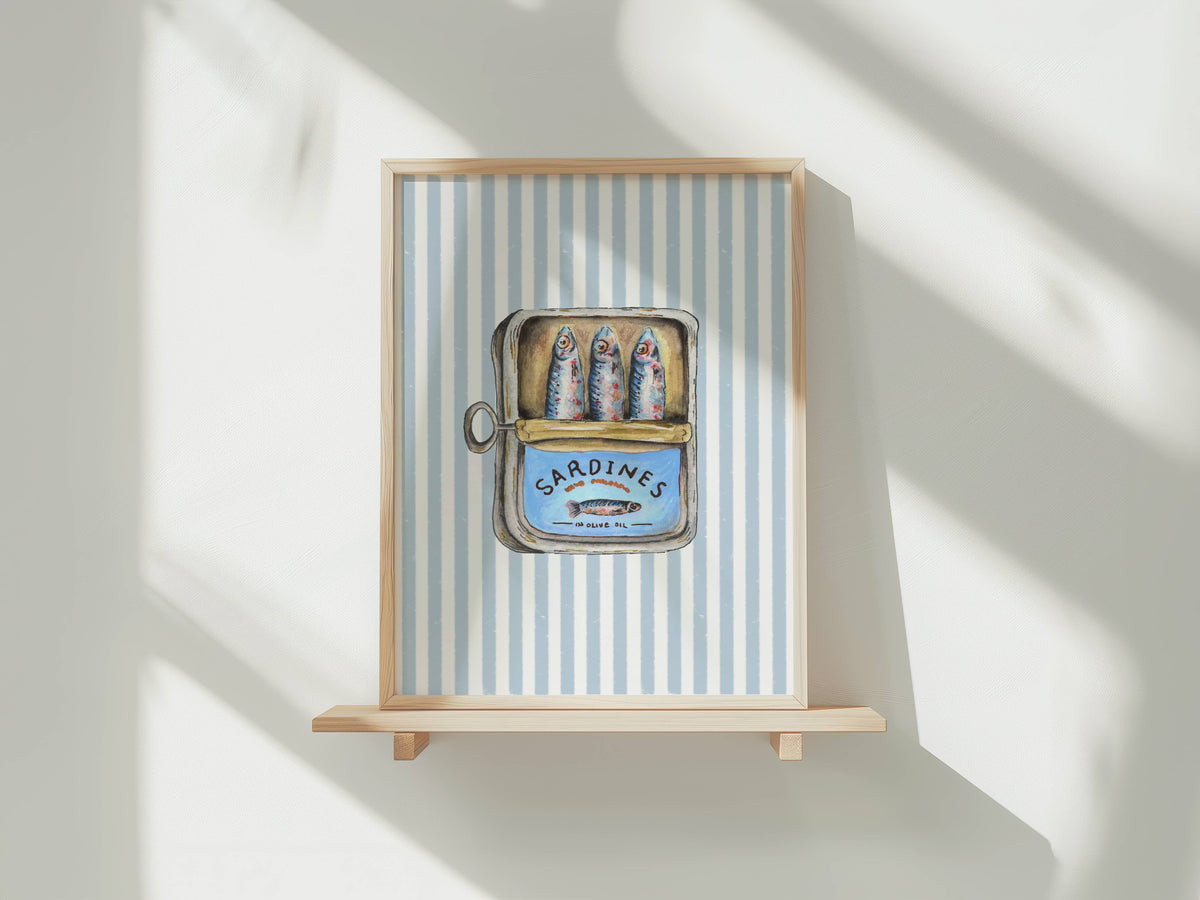 Framed wall art hanging on a light wood shelf against a white wall, with soft window shadows cast across the scene. The artwork features a blue-and-white striped background and a central illustrated tin of sardines labeled 'SARDINES'.