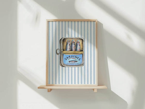 Framed wall art hanging on a light wood shelf against a white wall, with soft window shadows cast across the scene. The artwork features a blue-and-white striped background and a central illustrated tin of sardines labeled 'SARDINES'.