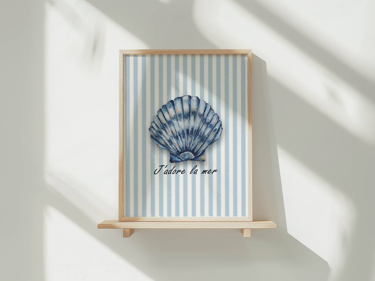 A framed art print featuring a blue and white striped background with a blue seashell in the center and the French text 'J'adore la mer' below it, displayed on a light wooden shelf against a white wall with soft window shadows.