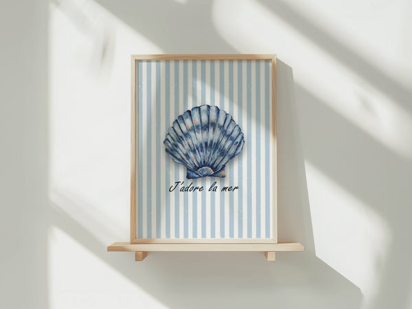 A framed art print featuring a blue and white striped background with a blue seashell in the center and the French text 'J'adore la mer' below it, displayed on a light wooden shelf against a white wall with soft window shadows.