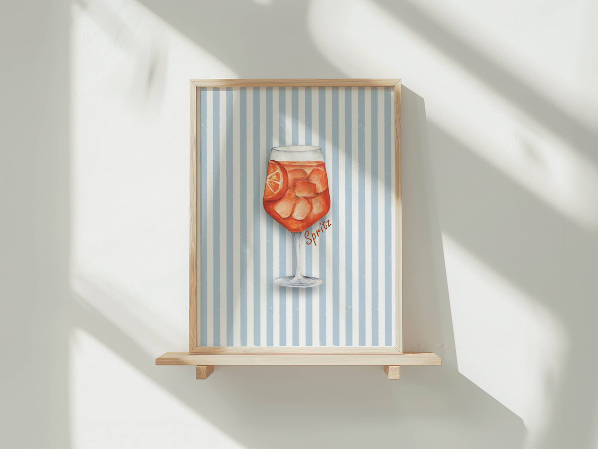 A framed art print featuring a watercolor illustration of an orange cocktail on a light blue and white striped background, displayed on a wooden shelf against a white wall with soft window shadows.