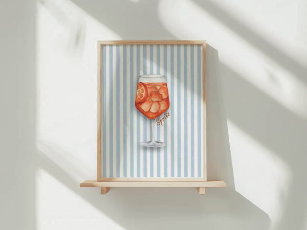 A framed art print featuring a watercolor illustration of an orange cocktail on a light blue and white striped background, displayed on a wooden shelf against a white wall with soft window shadows.