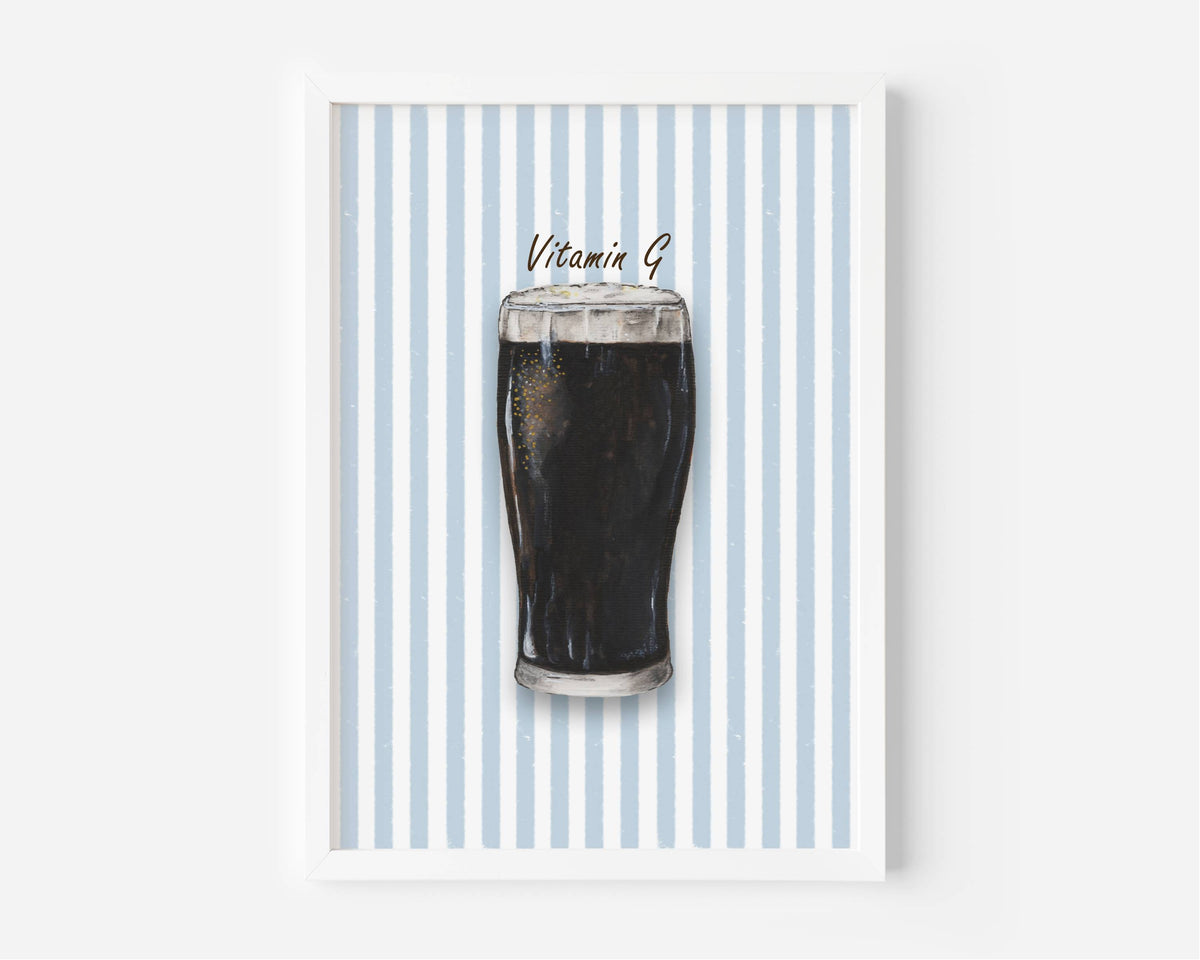 A framed art print featuring a dark pint of stout on a background of vertical light blue and white stripes, with the text 'Vitamin B' written above the drink. The print is housed in a clean white frame and is displayed against a neutral light gray wall.