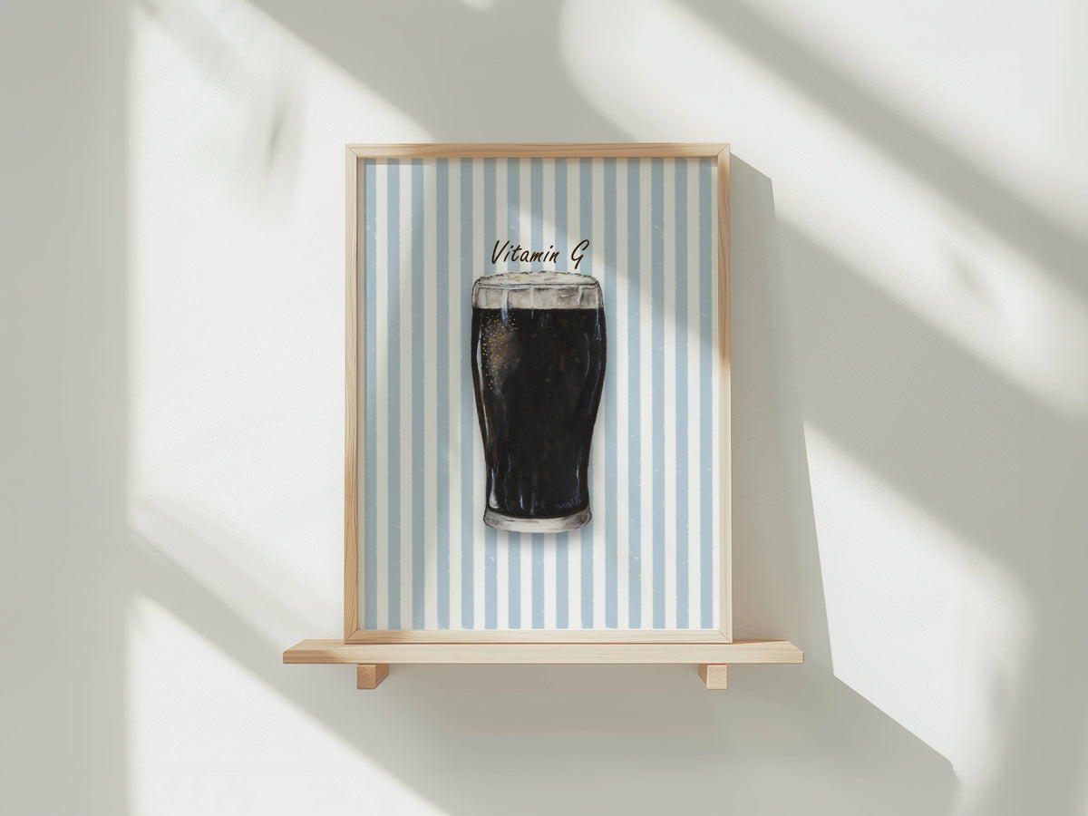 A framed art print featuring a pint of dark stout with 'Vitamin B' written above it, displayed on a light wooden shelf against a white wall with soft window shadows.