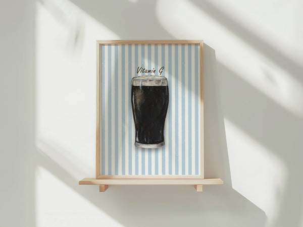 A framed art print featuring a pint of dark stout with 'Vitamin B' written above it, displayed on a light wooden shelf against a white wall with soft window shadows.