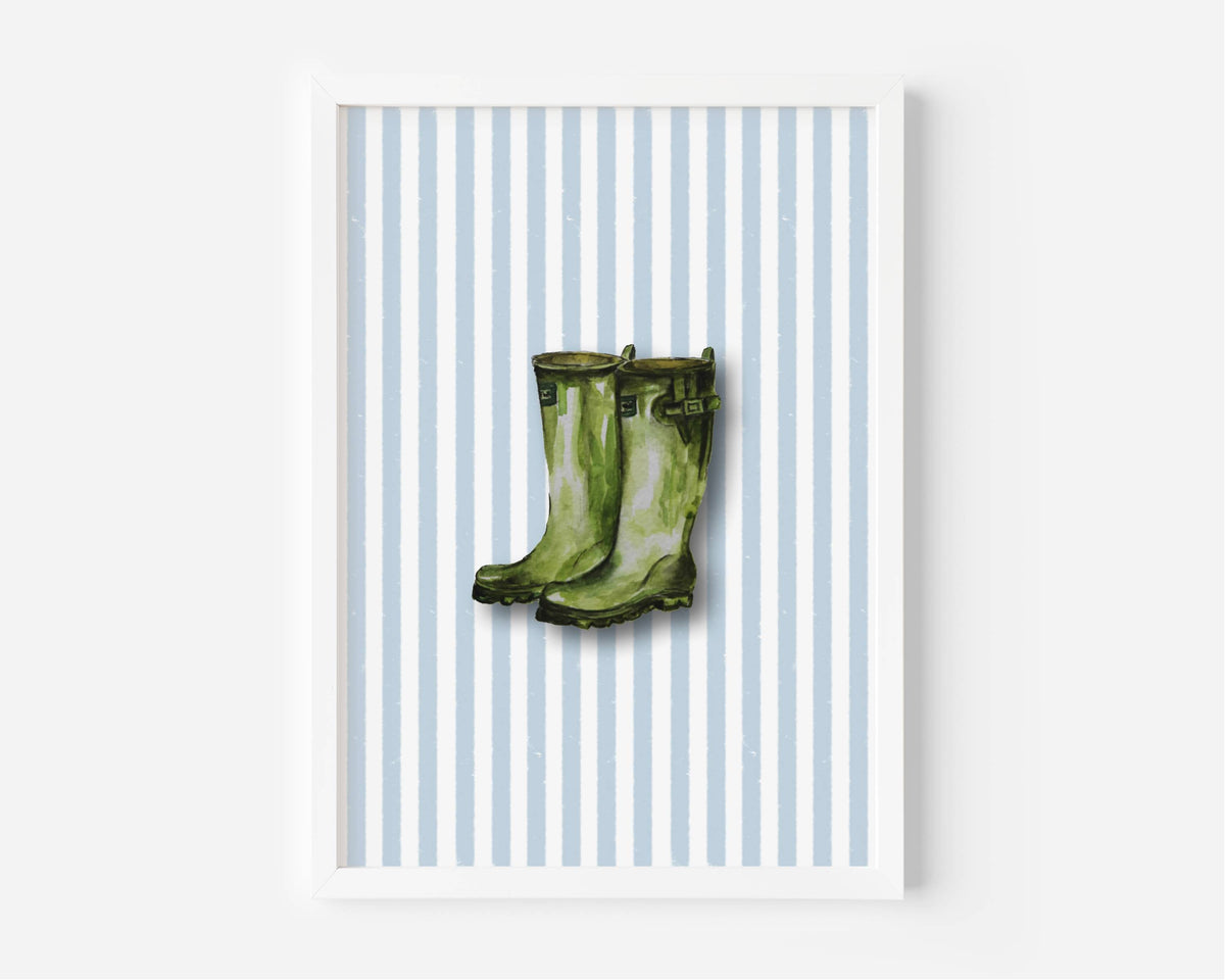 A framed art print featuring a pair of green rain boots centered on a background of light blue and white vertical stripes, displayed against a clean white wall.