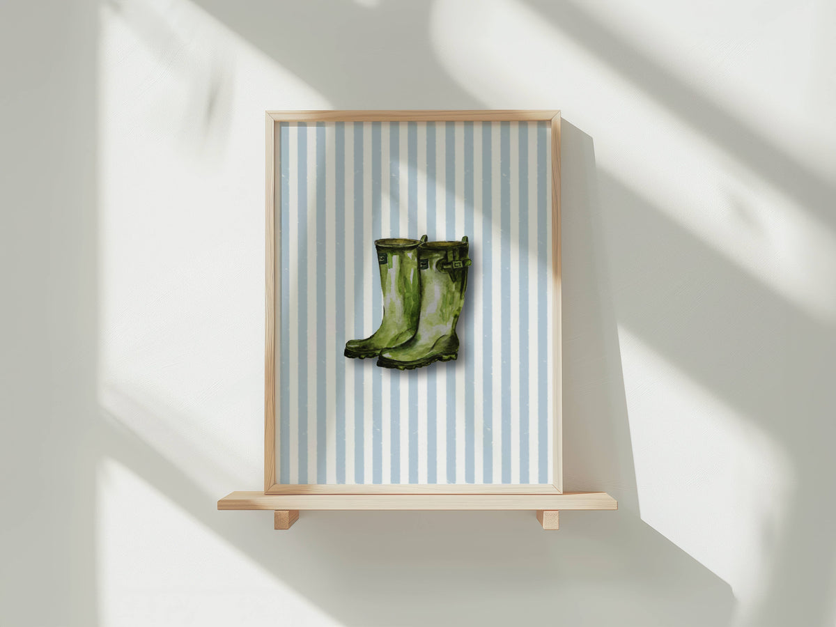 A framed art print featuring a pair of green rain boots on a light blue and white striped background, displayed on a light wood floating shelf against a white wall with soft window shadows.